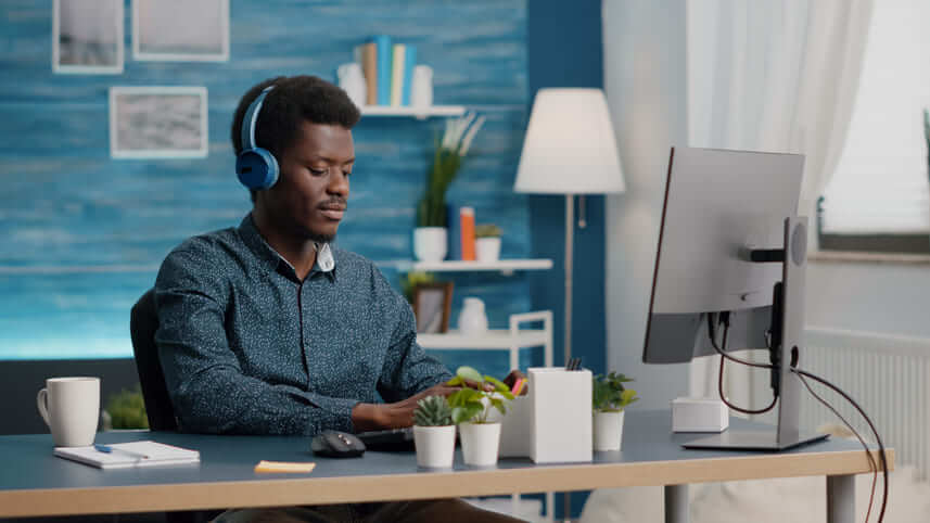 Programador freelancer: homem negro trabalhando em uma mesa com computador, usando fone, com parede azul ao fundo